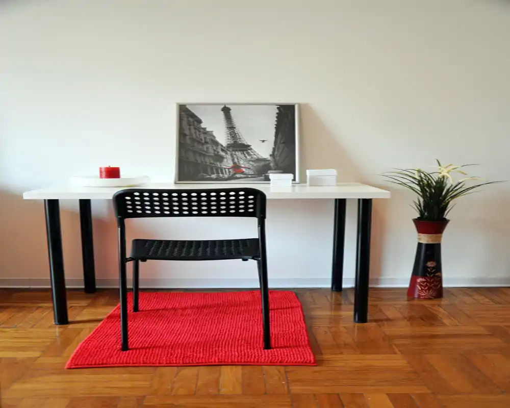 Spazio di lavoro compatto e minimal con scrivania bianca, sedia nera traforata e tappeto rosso; stampa decorativa e pianta completano l’ambiente.