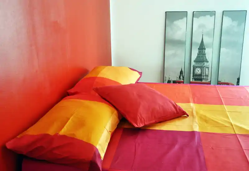 Camera da letto colorata con un grande letto rivestito da biancheria rossa e arancione, arricchito da cuscini e quadri. Scatto luminoso e ben composto, ideale per annunci che evidenziano un angolo notte accogliente e moderno.