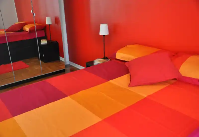 Camera da letto vivace con un ampio letto rosso-arancione che occupa la maggior parte dell’inquadratura, affiancato da una lampada da comodino e da un armadio con ante a specchio che danno profondità.