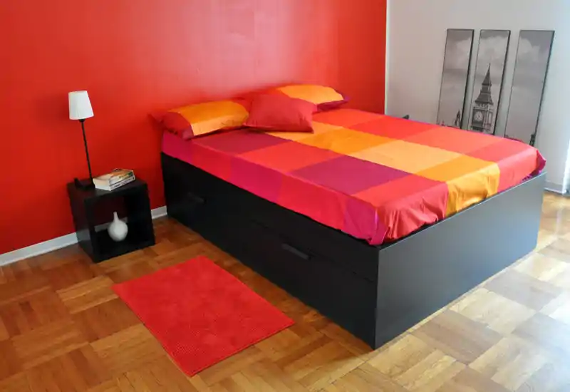 Camera da letto luminosa e moderna con un ampio letto con biancheria colorata, comodino con lampada e pavimento in parquet caldo. Linee pulite e una parete accentuata rossa creano un’atmosfera contemporanea e accogliente, ideale per annunci.