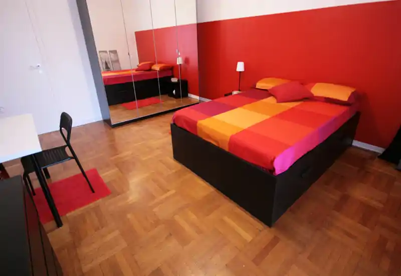 Camera da letto luminosa e ordinata con letto matrimoniale con biancheria rossa e arancione, armadio con ante specchiate, pavimento in parquet e lampada da comodino — accogliente e perfetta per annunci.