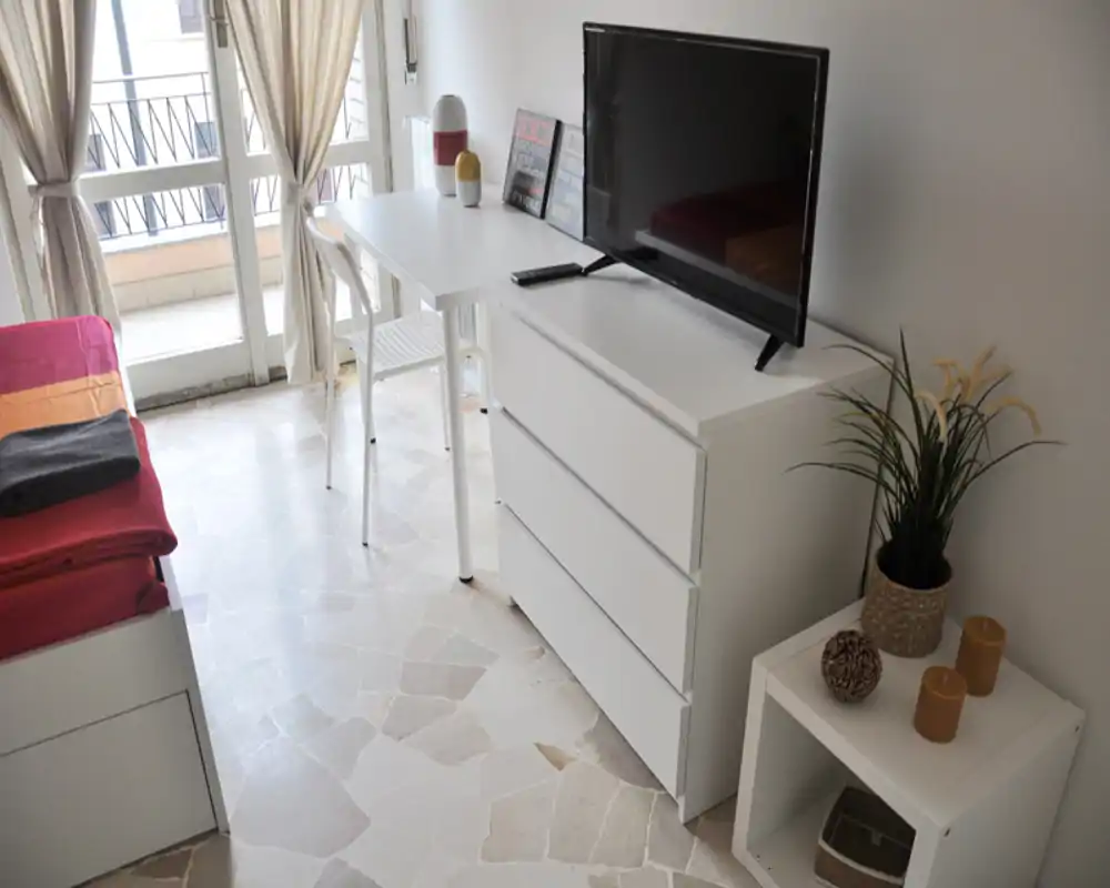 Angolo camera da letto luminoso e moderno con letto singolo, comò bianco con TV, un piccolo tavolino con oggetti decorativi e una scrivania vicino alla porta sul balcone che lascia entrare luce naturale.