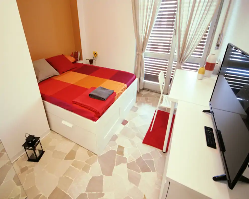 Camera da letto luminosa e accogliente con letto matrimoniale con copriletto rosso e arancione, scrivania vicino alla finestra e TV su un mobile basso. La luce naturale entra dalle tende e il pavimento piastrellato aggiunge carattere.