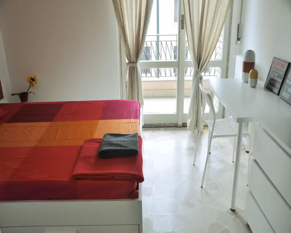Camera da letto luminosa e ben curata con letto colorato, scrivania e accesso al balcone; luce naturale abbondante.
