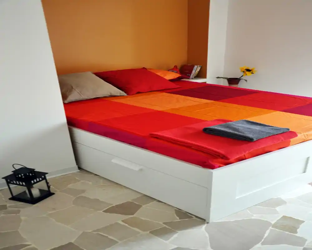 Camera da letto luminosa e ordinata con copriletto sui toni del rosso e arancione, cuscini e vano contenitore sotto il letto; parete accentuata calda e decorazioni semplici per un ambiente accogliente e moderno.