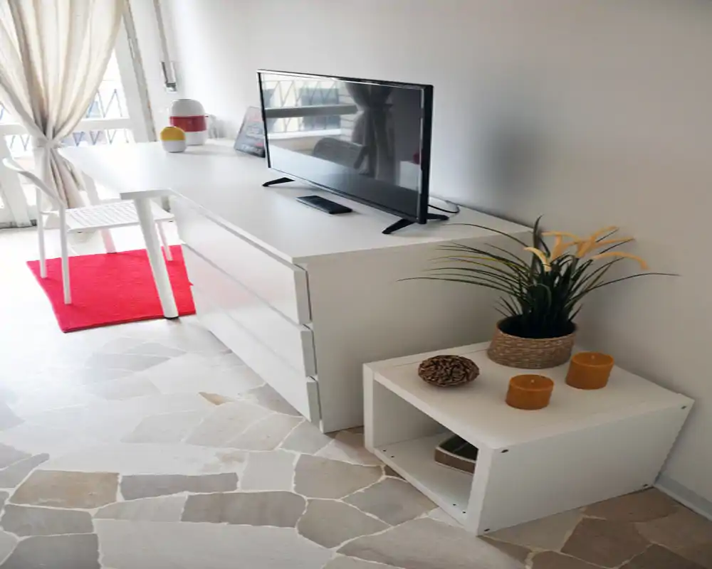 Angolo soggiorno luminoso e minimal con TV su un comò bianco, pianta e candele decorative, e luce naturale dalla finestra/porta balcone.
