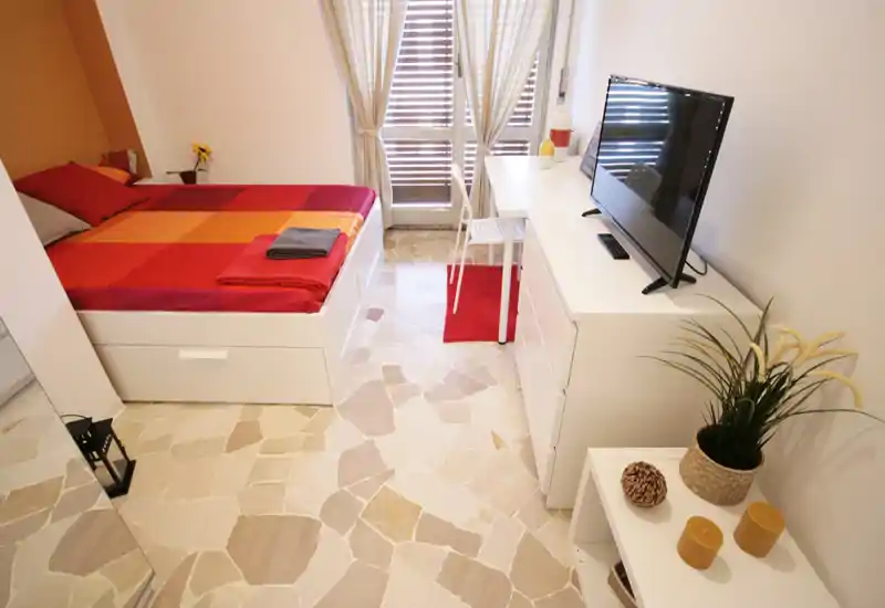 Camera da letto luminosa e curata con un letto matrimoniale colorato, scrivania vicino alla finestra e TV su un comò basso — ideale per una persona o una coppia in cerca di un soggiorno accogliente in città.