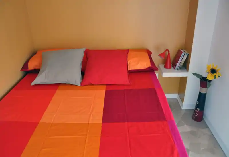 Camera da letto luminosa e colorata con un letto matrimoniale coperto da biancheria rossa e arancione, due cuscini e un semplice comodino a mensola con lampada e libri.