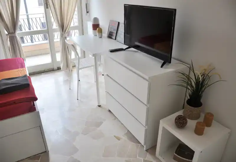 Bright, modern bedroom corner with a single bed, white dresser topped by a TV, a small side table with decor and a desk by a balcony door letting in natural light.