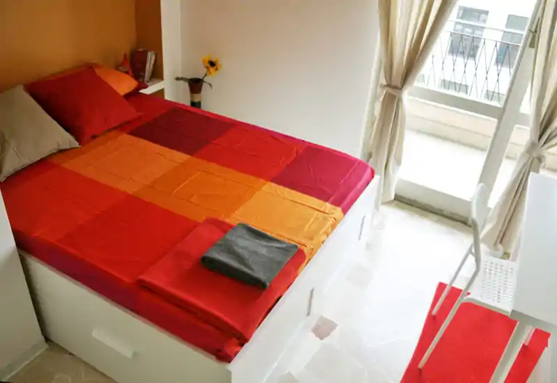 Camera da letto luminosa e accogliente con un letto matrimoniale dai colori vivaci vicino a una finestra che lascia entrare molta luce naturale; scrivania e sedia semplici per un layout compatto e funzionale.