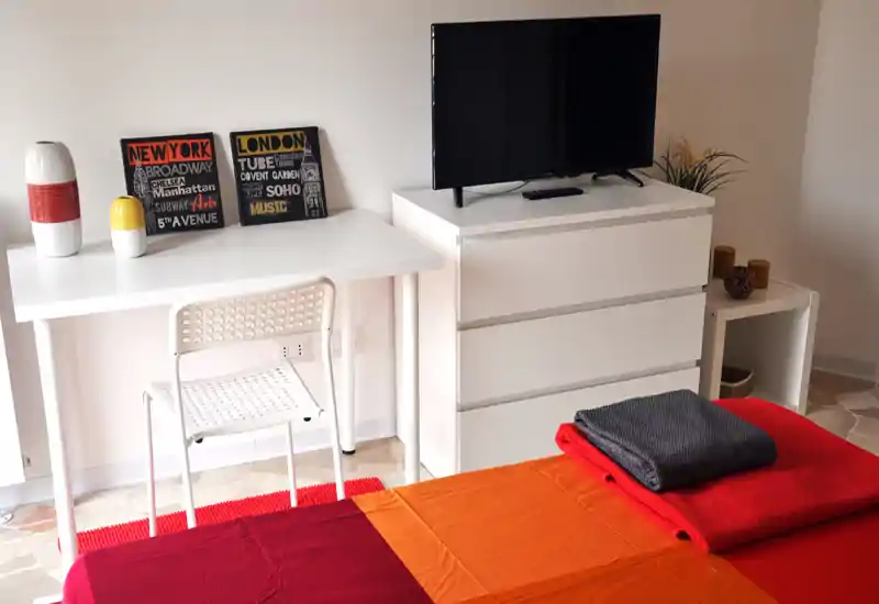 Camera da letto luminosa e ben arredata con una coperta vivace, plaid piegati e una scrivania; la TV è posizionata su un comò bianco, ambiente ordinato e accogliente.