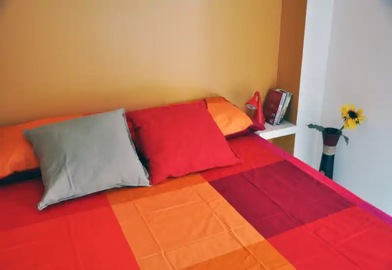 Camera da letto luminosa e accogliente con copriletto rosso-arancione, cuscini e un piccolo ripiano con lampada e libri.