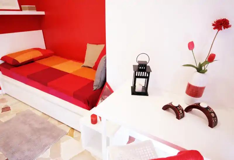 Bright, stylish single bedroom with a red accent wall and a colorful checked bedspread, small desk area with decorative vase and lantern — ideal for a cozy single stay.