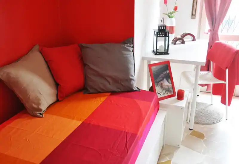 Compact, brightly styled bedroom corner with a red accent wall, colorful bedspread and decorative cushions; a small desk by the window brings natural light and a cozy workspace.