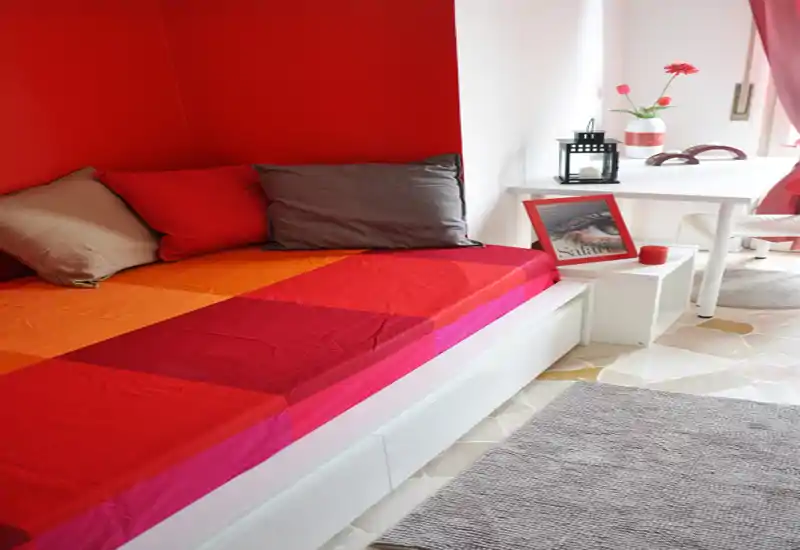 Bright, colorful bedroom corner with a single bed dressed in warm red and orange linens, decorative cushions, and a small workspace by the window.