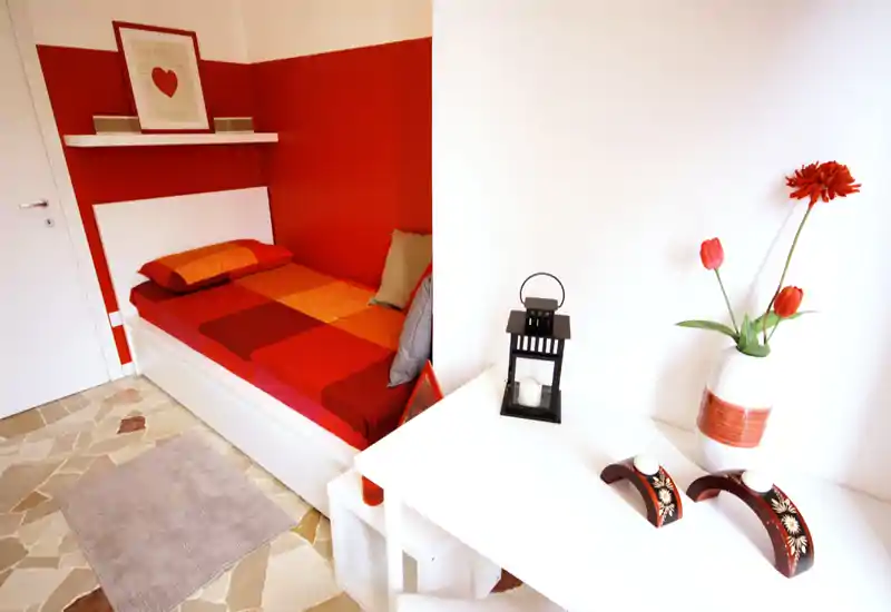 Compact single bedroom with a colorful red-and-orange bed, white furniture and decorative accents; bright, well-styled and welcoming space ideal for a short-stay rental.