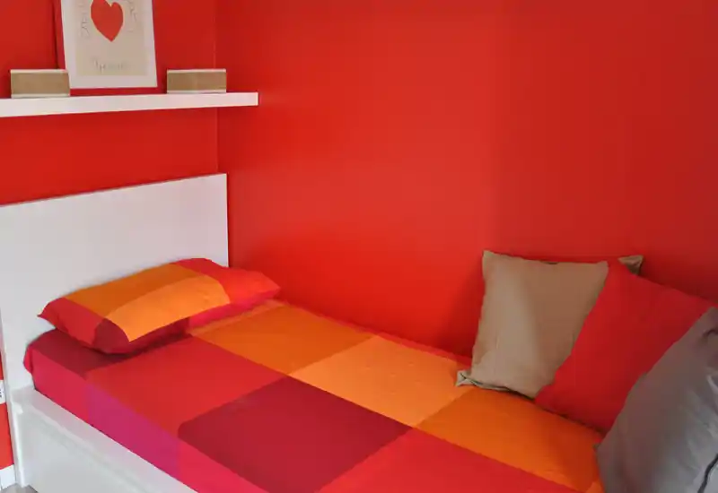 Camera da letto compatta e luminosa con un letto singolo coperto da biancheria rossa e arancione e cuscini decorativi; testiera bianca e mensola che creano un ambiente ordinato e accogliente.