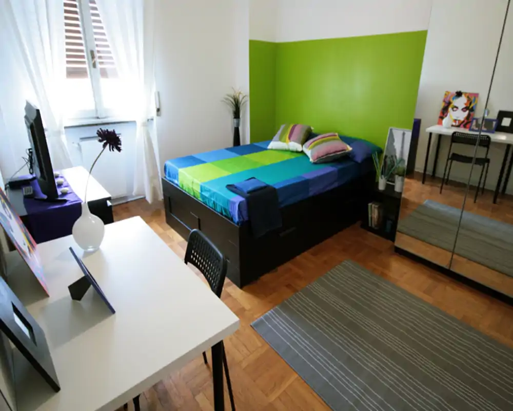 Camera da letto luminosa e moderna con letto matrimoniale, biancheria colorata, contenitori sotto il letto e armadio con ante a specchio che amplia la stanza. Scrivania vicino alla finestra e decorazioni sobrie rendono l'ambiente funzionale e accogliente.