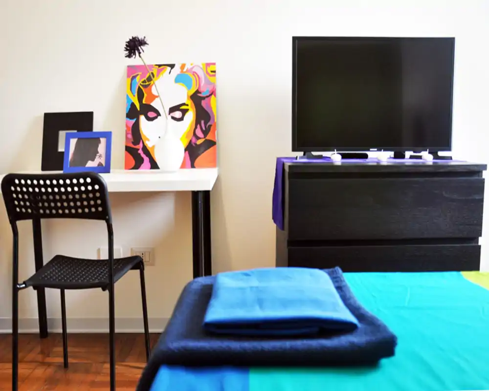 Camera da letto moderna e luminosa: asciugamani piegati sul letto in primo piano, una scrivania semplice con sedia e una TV su comò scuro. Linee pulite, un’opera colorata e tessili ordinati rendono l’ambiente accogliente.