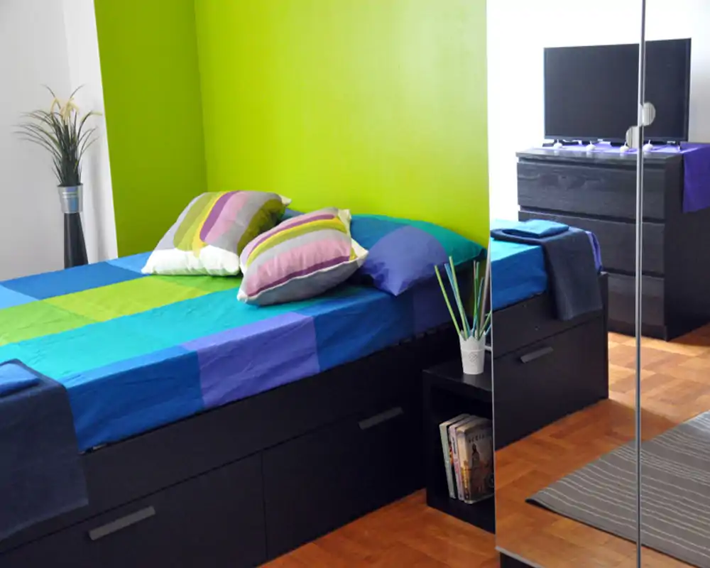 Camera da letto luminosa e colorata con letto matrimoniale ben rifatto, cuscini decorativi e base contenitore nera moderna. Armadio con specchio, parquet e comò con TV completano uno spazio compatto e curato, perfetto per annunci.