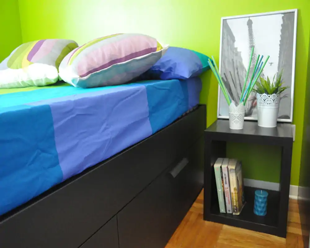 Angolo di camera luminoso e colorato con un letto ben rifatto con cassetti sotto e un comodino con decorazioni e libri — area notte compatta e ben arredata.