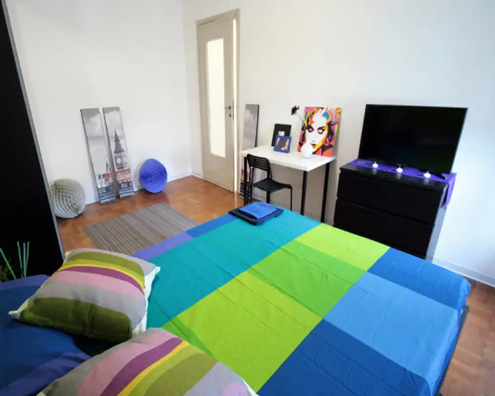 Camera da letto luminosa e colorata con un ampio letto in primo piano, cuscini a righe e biancheria sistemata, con scrivania e TV sulla parete opposta.