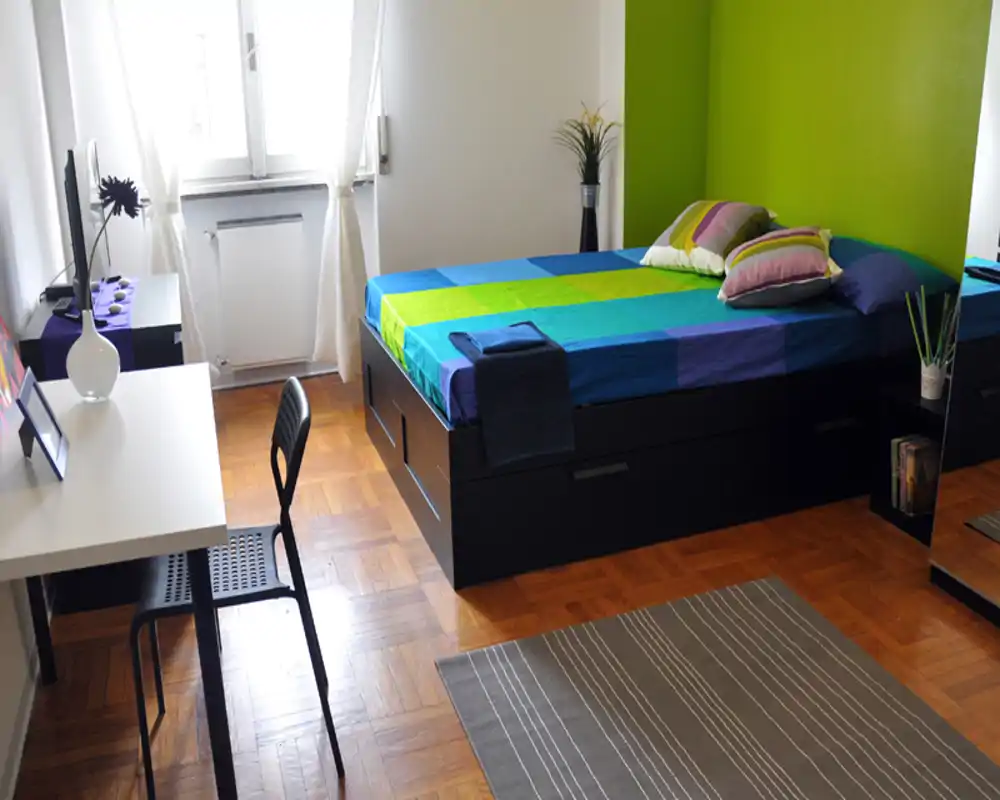 Camera da letto singola luminosa e ben arredata con letto colorato, scrivania e luce naturale dalla finestra — ordinata e pronta per un annuncio.