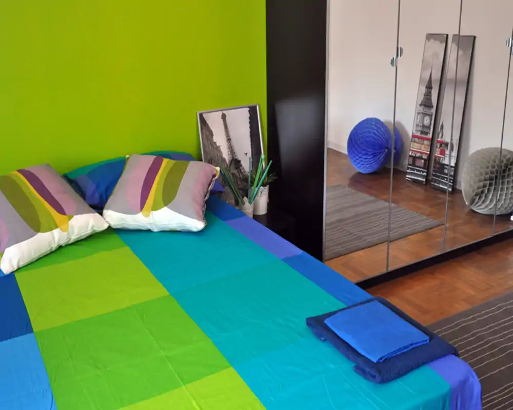 Camera da letto luminosa e colorata con un ampio letto coperto da biancheria blu e verde, cuscini decorativi e armadi con ante a specchio che ampliano la stanza.