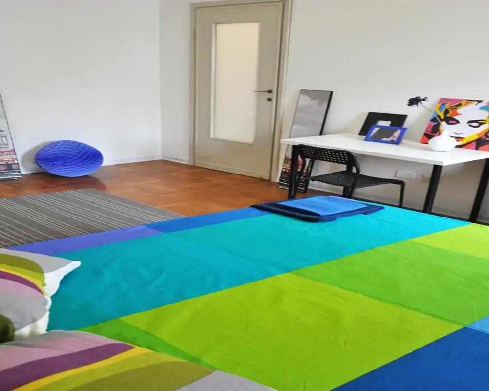 Camera da letto luminosa e colorata con un grande letto coperto da una trapunta a righe vivaci. L'ambiente è arredato con semplicità: scrivania, sedia, quadri e pavimenti in legno che rendono lo spazio accogliente e ideale per affitti brevi.