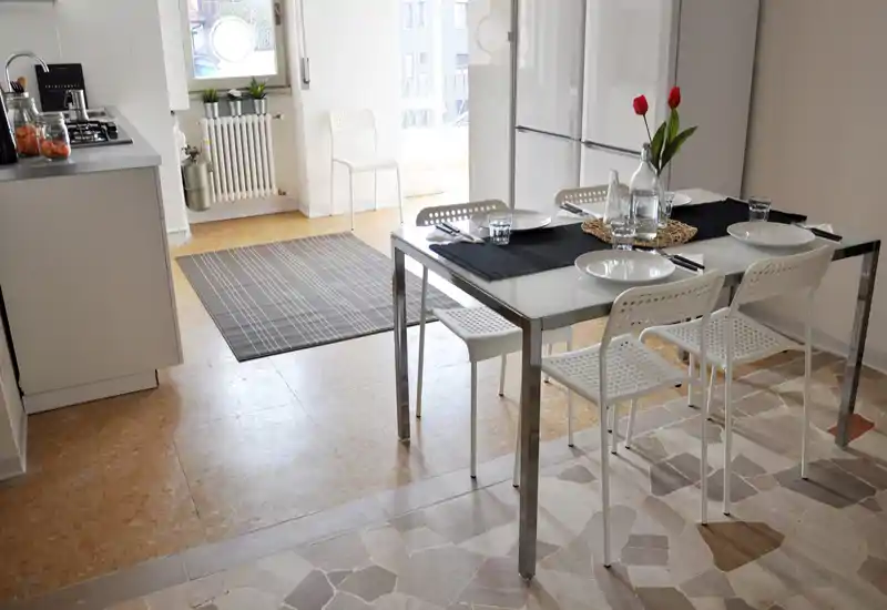 Bright, modern dining area adjacent to the kitchen with a glass-top table set for four and natural light from a balcony/large window.