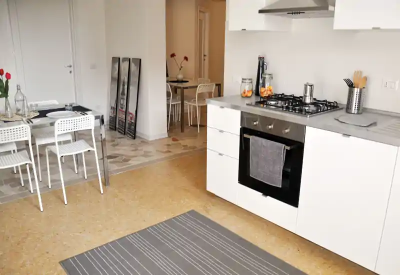 Modern open kitchen with white cabinets, stainless-steel gas hob and built-in oven; adjacent dining area with a small table and white chairs, decorated with simple vases and a grey striped rug.