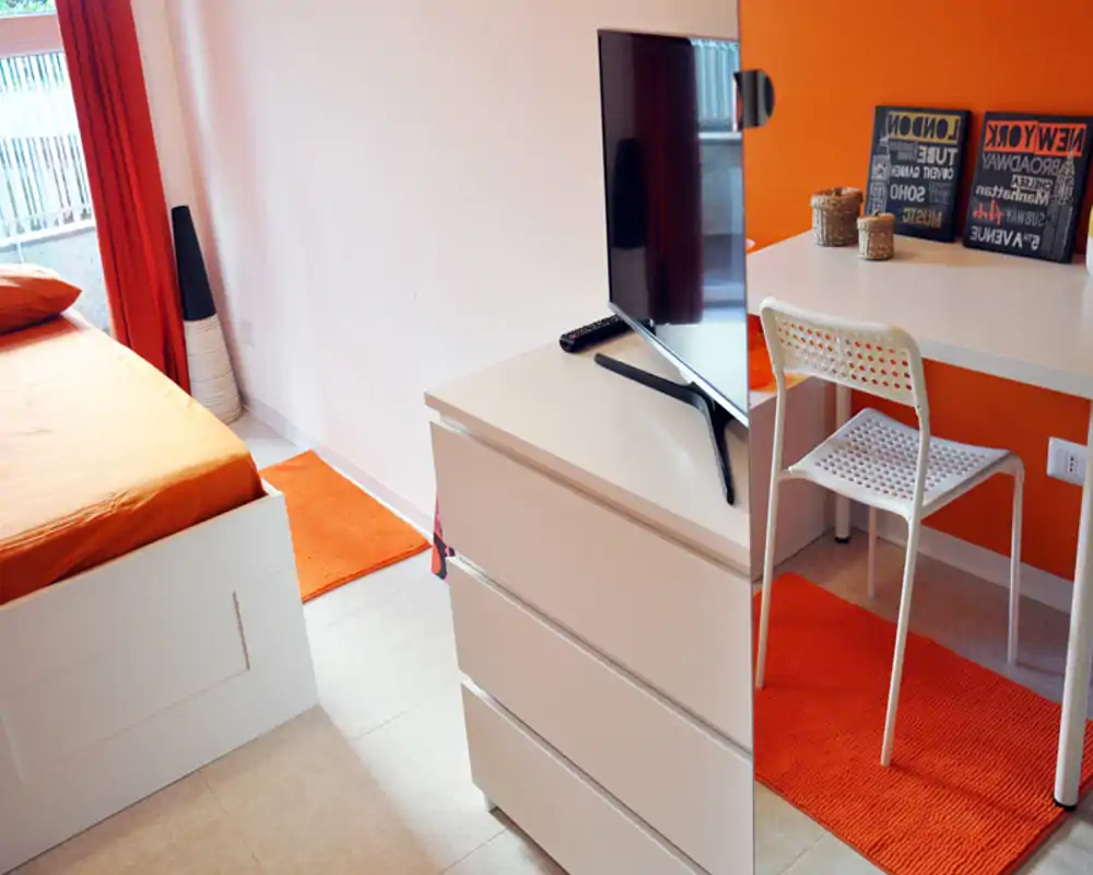 Camera da letto luminosa e ben arredata con accenti arancioni, letto visibile, comò e un piccolo angolo scrivania — accogliente e moderna.