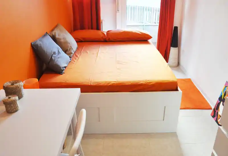 Camera da letto moderna e luminosa con un grande letto rivestito di arancione che occupa la maggior parte della stanza, arricchito da tessuti caldi e luce naturale dalla finestra.