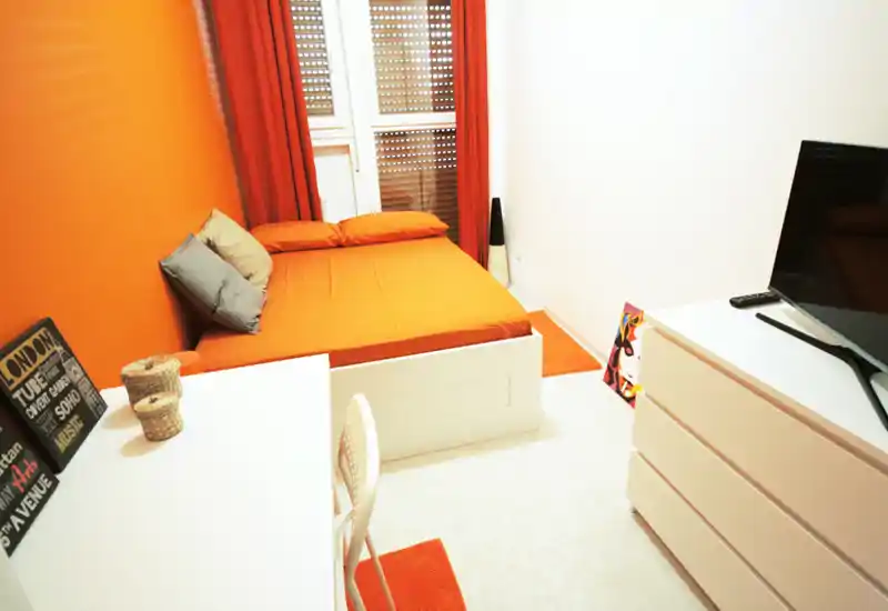 Camera da letto compatta e luminosa con arredi moderni, letto matrimoniale, scrivania e comò; tonalità arancioni calde e buona luce naturale.