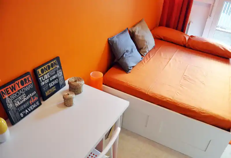 Camera da letto compatta e luminosa con parete arancione e letto matrimoniale ben rifatto. Un piccolo scrittoio con decorazioni e cesti è accanto al letto e una finestra garantisce luce naturale.