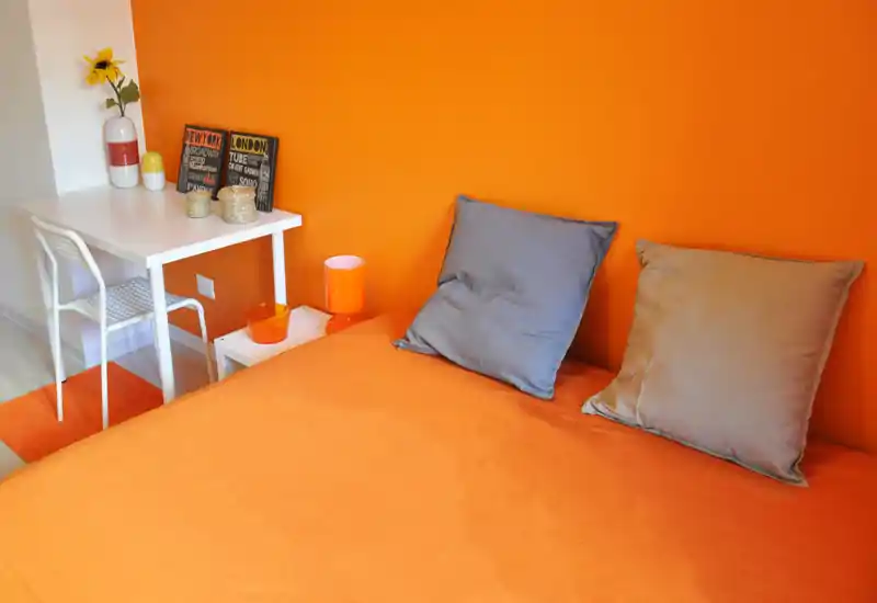 Camera da letto luminosa e moderna con parete arancione vivace e biancheria coordinata, due cuscini, comodino con lampada e una piccola scrivania — ambiente accogliente e colorato ideale per affitti brevi.