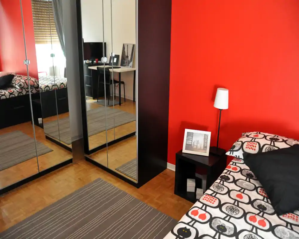 Dormitorio moderno y luminoso con pared roja de acento, armario con puertas espejo y ropa de cama estampada. Mesita con lámpara y foto decorativa que aportan confort.