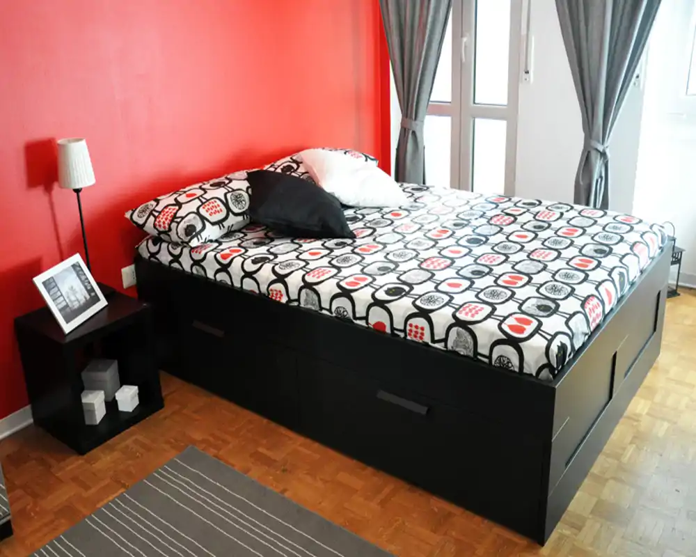 Dormitorio luminoso y bien arreglado con una amplia cama plataforma, ropa de cama estampada y pared de acento roja. La luz natural desde la ventana y los muebles simples crean un ambiente moderno y acogedor.