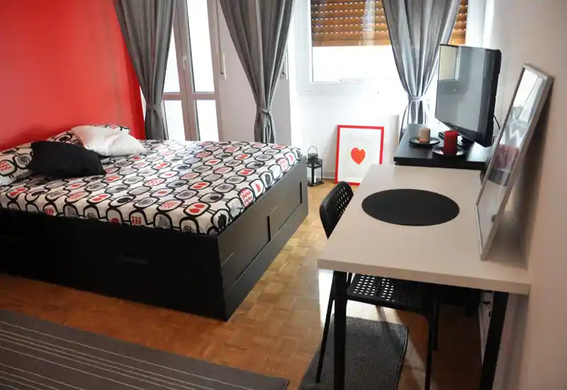 Camera da letto moderna e luminosa con letto matrimoniale con cassettoni sottostanti, scrivania e TV vicino alla finestra. Tende grigie e parete rossa come accento donano carattere, ideale per un annuncio di locazione.