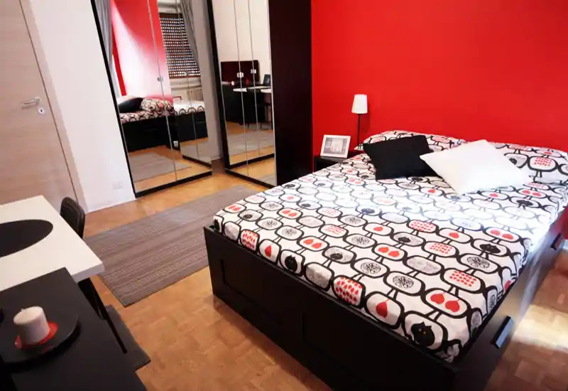 Camera da letto moderna e luminosa con un ampio letto matrimoniale, biancheria stampata e armadi a specchio. La parete rossa e il pavimento in parquet creano un'atmosfera calda e di design.