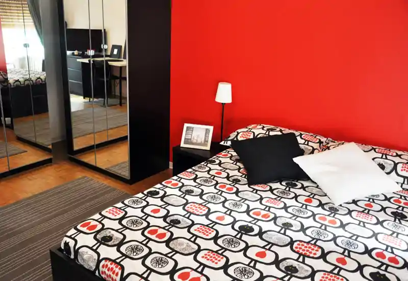 Camera da letto luminosa e moderna con copriletto fantasia, cuscini decorativi e una parete rossa d'accento; armadio con specchi e pavimento in legno che ampliano lo spazio.