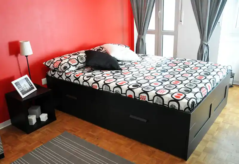 Camera da letto luminosa e ben curata con grande letto piattaforma, biancheria a fantasia e parete d'accento rossa. La luce naturale dalla finestra e il semplice comodino creano un'atmosfera moderna e accogliente.