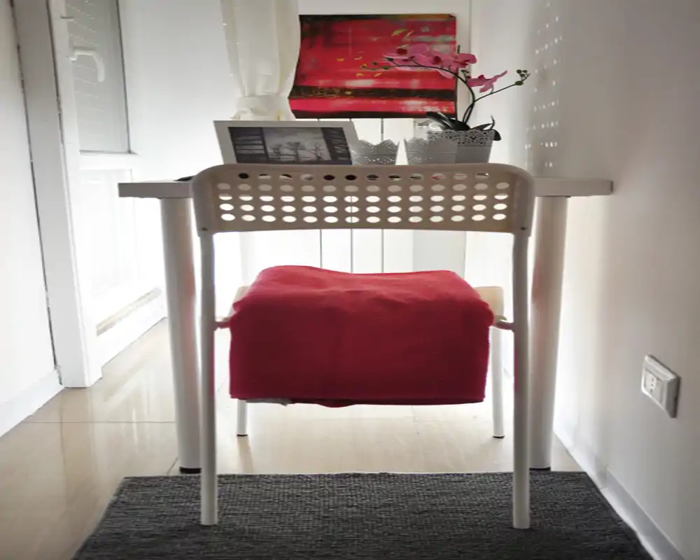 Bright, narrow seating or workspace corner featuring a simple desk, perforated chair and decorative accents (red throw, small plant and framed print). Ideal for a cozy single-person nook in a living area or studio.