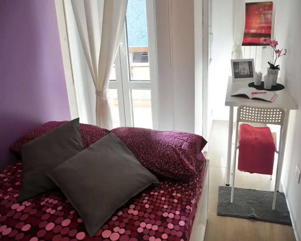 Bright, cozy bedroom with patterned bedding, pillows and a small desk by a sunlit window — an inviting, well-lit space ideal for a single occupant.