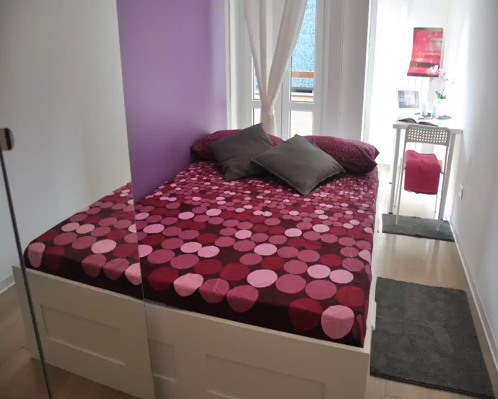 Bright, well-styled bedroom with a large double bed covered in patterned bedding and a small desk by the window, ideal for a cozy stay.