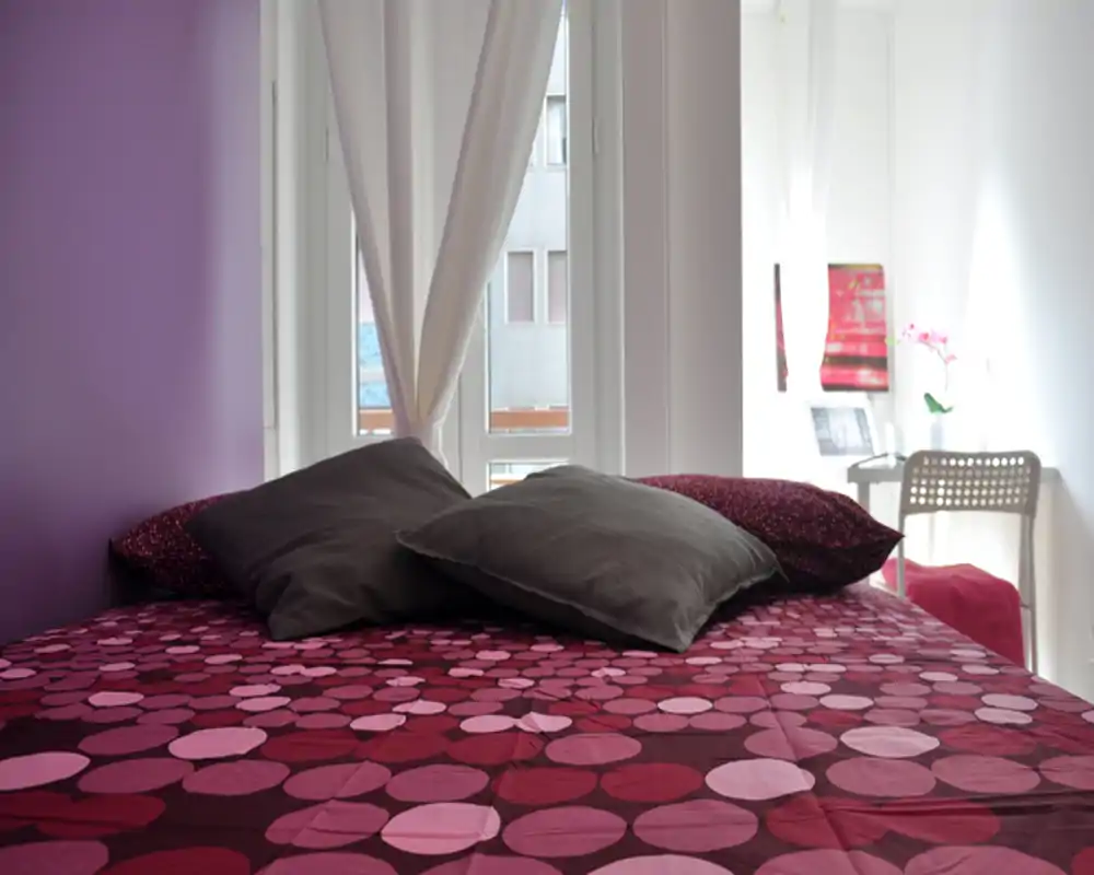 Bright, cozy bedroom with a patterned plum duvet and multiple pillows facing a window; natural light and modern accents create a welcoming atmosphere.
