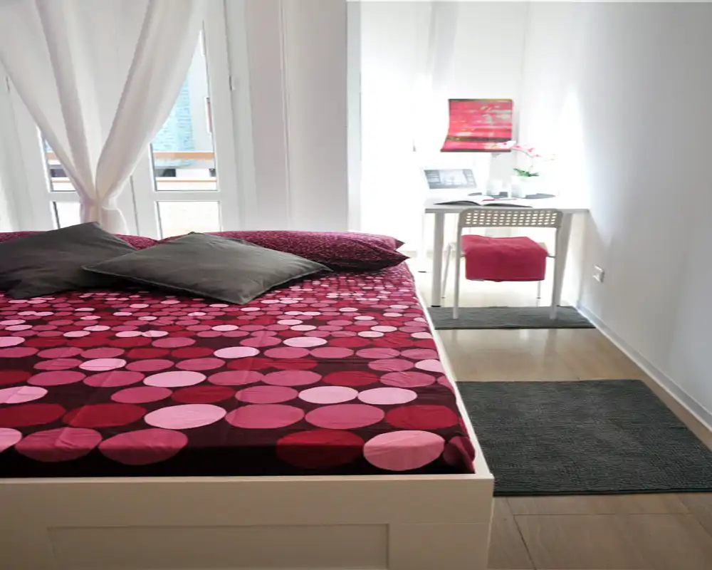 Bright, well-styled bedroom with a patterned double bed, cushions and a small desk by the window — ideal for a cozy city stay.