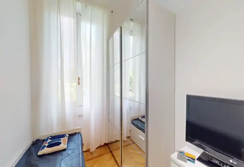 Small bright bedroom with a single daybed by a large window, sheer curtains and a mirrored wardrobe reflecting light. Clean parquet floor and a TV on a small cabinet complete the compact, functional layout.
