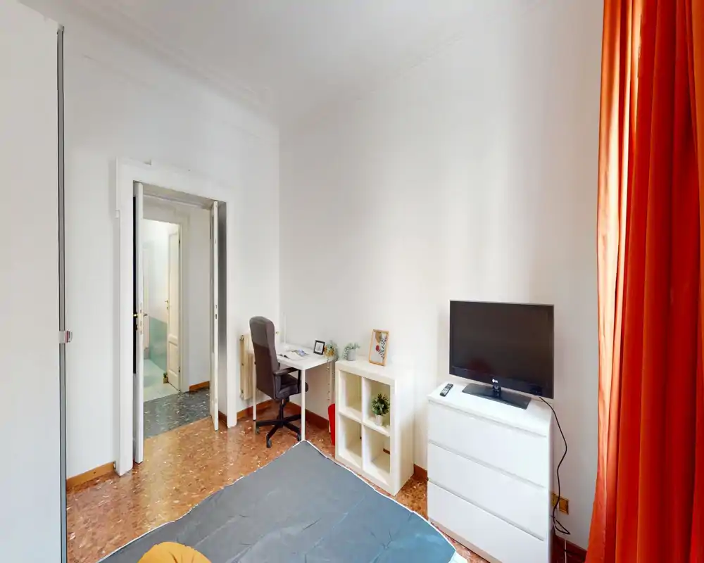 Bright, simply furnished bedroom with a visible bed, desk and TV. The room features warm orange curtains and a compact workspace beside shelving and a chest of drawers.