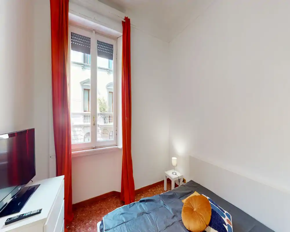 Bright, tidy bedroom with a double bed, warm red curtains and a bedside lamp; natural light from a tall window enhances the room.